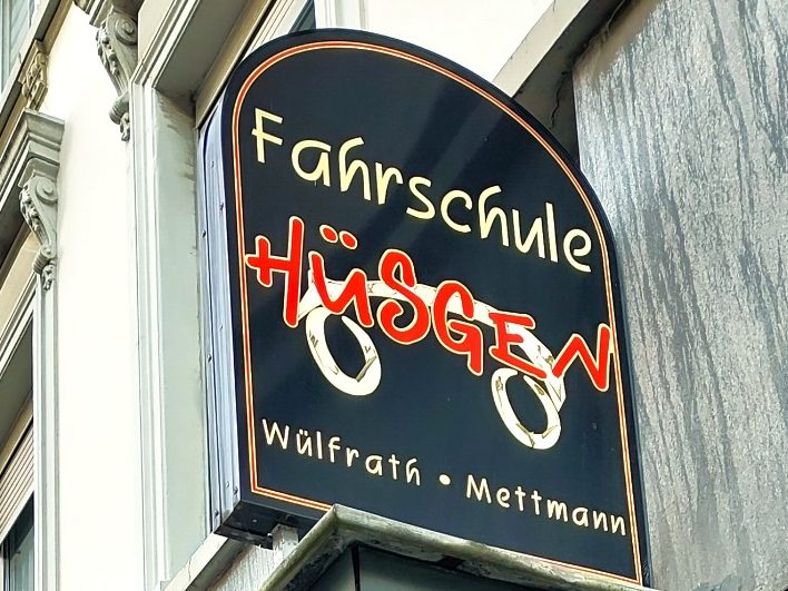 Schwarzes Schild mit der Aufschrift "Fahrschule Hüsgen" in roten Buchstaben.