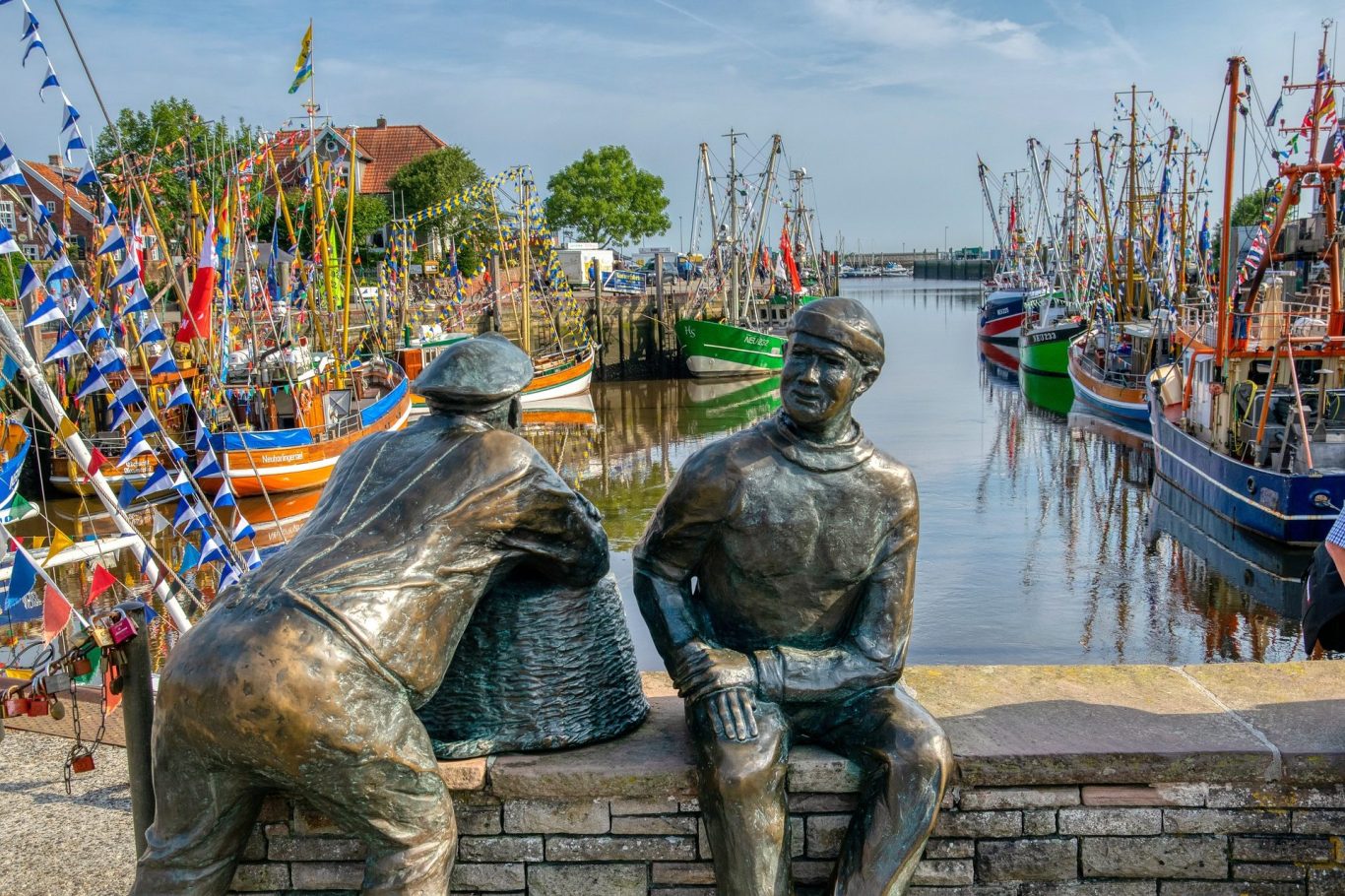 Bronzeskulpturen von zwei Männern am Wasser, umgeben von festlich geschmückten Booten.