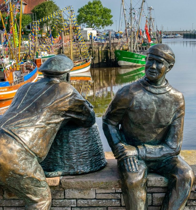 Zwei bronzene Figuren am Hafen, eine lehnt sich an eine Kiste, die andere sitzt entspannt.